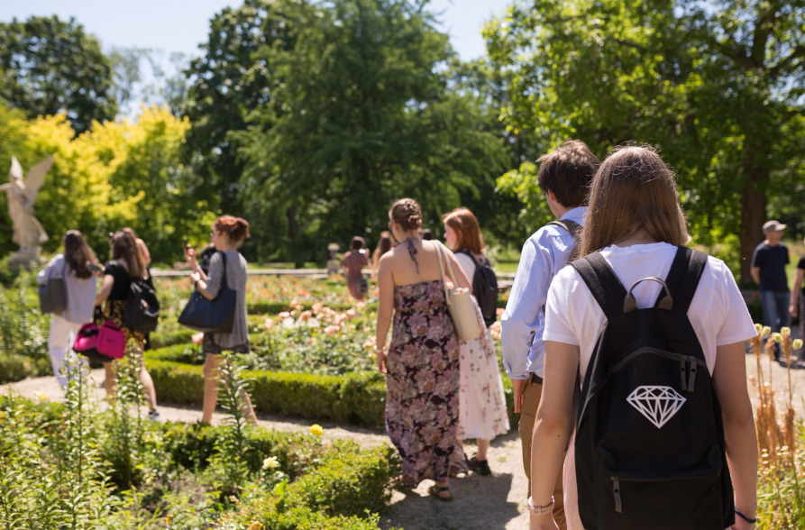 Kolorowe zdjęcie współczesne. Przedstawia grupę osób spacerujących po alejkach w ogrodzie Pałacu w Wilanowie.