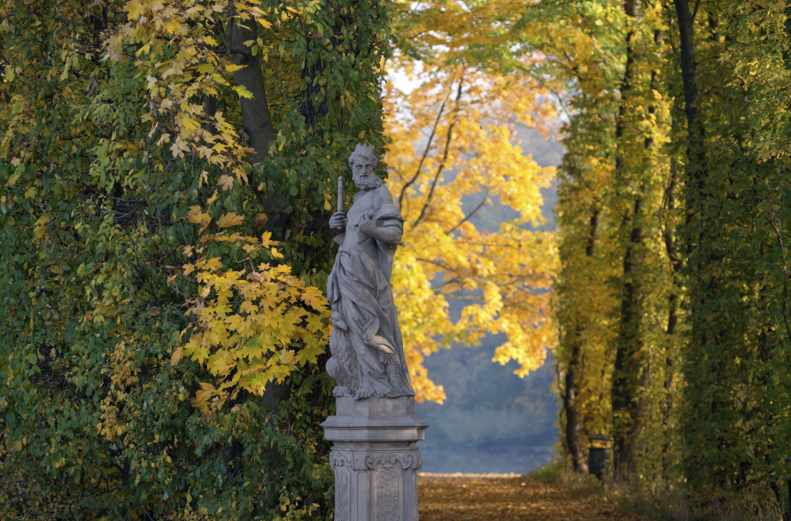 Kolorowe zdjęcie współczesne. Przedstawia rzeźbę na cokole na tle jesiennego pejzażu. Rzeźba to stojący mężczyzna z brodą, w długich szatach, trzymający w ręce przedmiot.