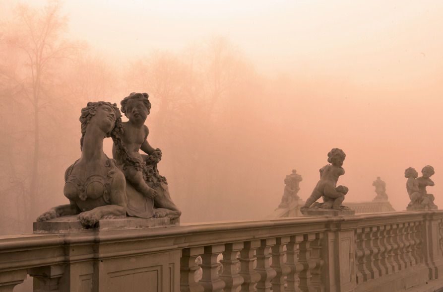 Kolorowe zdjęcie współczesne. Przedstawia balustradę i rzeźby. W tle zamglony widok na ogród.