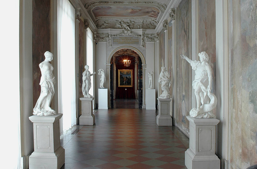 Kolorowa fotografia współczesna. Przedstawia widok na hol w pałacu wialnowskim. Po bokach stoją rzeźby antyczne na postymentach. Na ścianach z prawej strony freski.