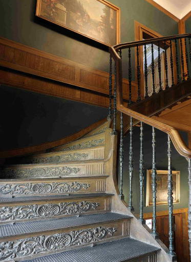 Kolorowe zdjęcie przesdtawia stare, bogato zdobione schody na klatce schodowej. Na ciemno-zielonych ścianach wiszą obrazy olejne.
