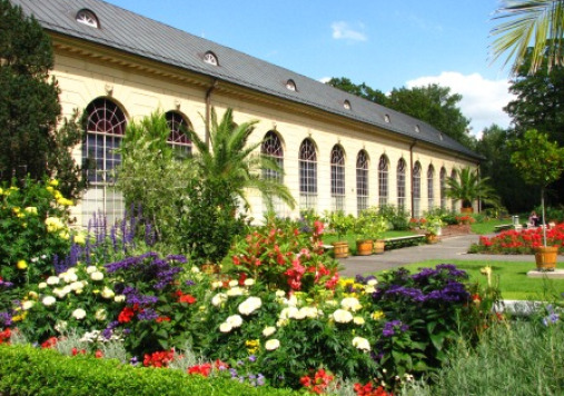 Kolorowe zdjęcie współczesne. Przedstawia niski budynek w otoczeniu kwiatów. Jest to oranżeria, którą zdobią rośliny egzotyczne.