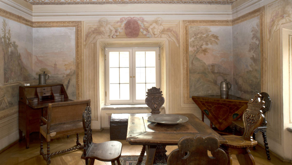 Kolorowe zdjęcie przedstawia fragment ekspozycji w Wilanowie. We wnętrzu ozdobionym freskami znajdują się meble i sprzęty antyczne.