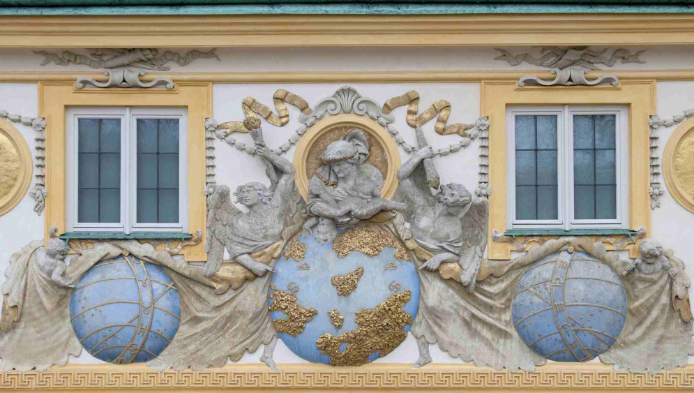 Zdjęcie współczesne. Przedstawia fragment elewacji pałacu w Wilanowie. Pomiędzy dwoma oknami płaskorzeźba - kula ziemska podtrzymywana przez dwa anioły i mężczyznę którego portret umieszczony jest w tondzie.