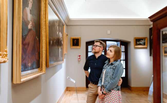 Kolorowe zdjęcie współczesne. Przedstawia parę: kobietę i mężczyznę, którzy stoją w skupieniu przed obrazem olejnym w galerii.