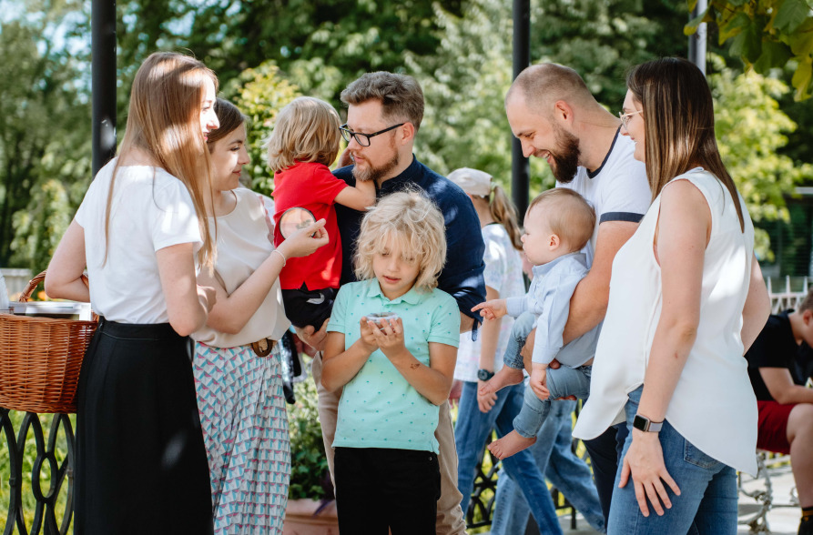 Kolorowe zdjęcie współczesne. Przedstawia grupę ludzi z dziećmi. Jest to spotkanie rodzin na tle pejzażu. Portretowani ubrani są w letnie stroje współczesne.