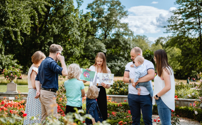 Kolorowe zdjęcie współczesne. Przedstawia dwie rodziny z dziećmi które stoją w parku. Przed grupą przewodniczka z książką w rękach, która coś im objaśnia.