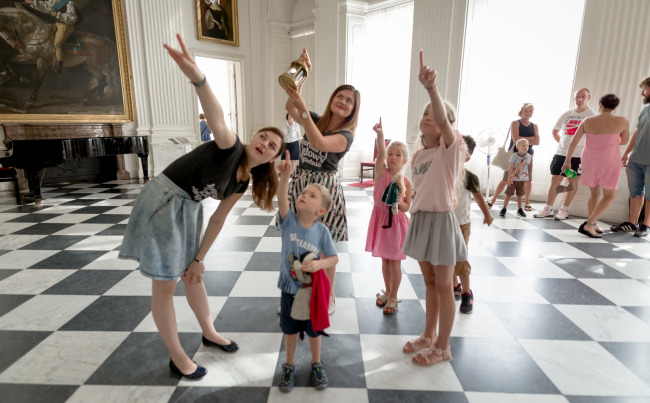 Kolorowe zdjęcie współczesne. We wnętrzu pałacu stoją dzieci z dwiema młodymi paniami, które pokazują im coś w górze.