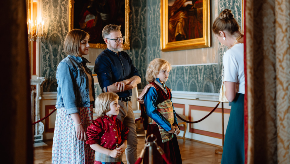 Kolorowe zdjęcie współczesne. We wnętrzu pałacu młodzi ludzie: kobieta i mężczyzna oraz dwóch małych chłopców. Jeden z nich ubrany jest w strój historyczny. Wszyscy zwróceni są w kierunku młodej kobiety, która im coś pokazuje.