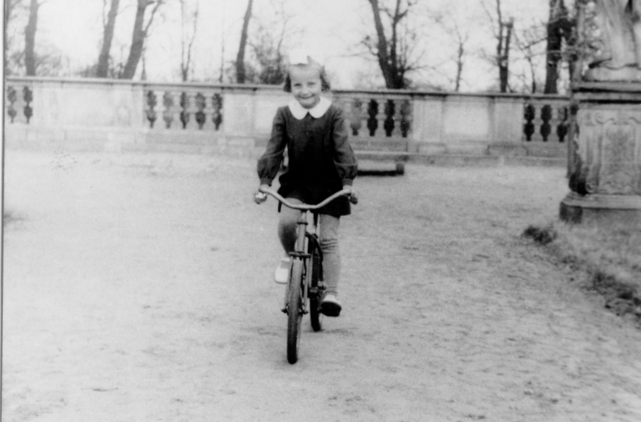 Dziedziniec pałacu. Uśmiechnięta dziewczynka jedzie rowerem.