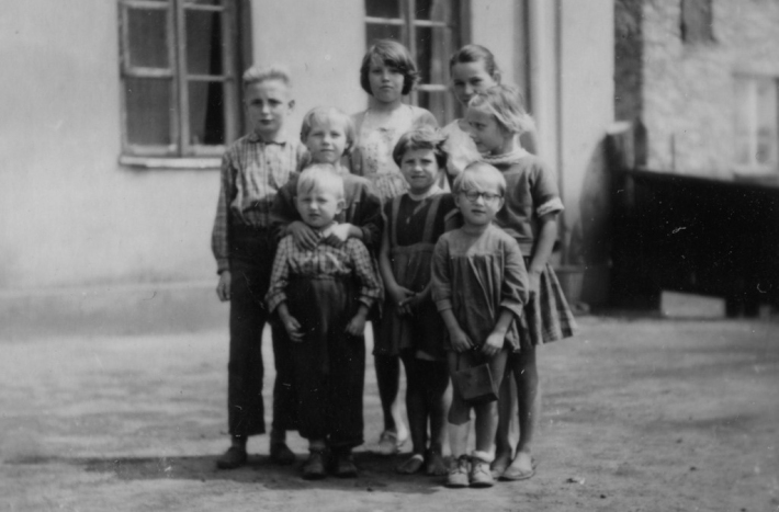 Grupa dzieci w różnym wieku pozuje na podwórku. Stoją wyprostowane i patrzą w obiektyw.