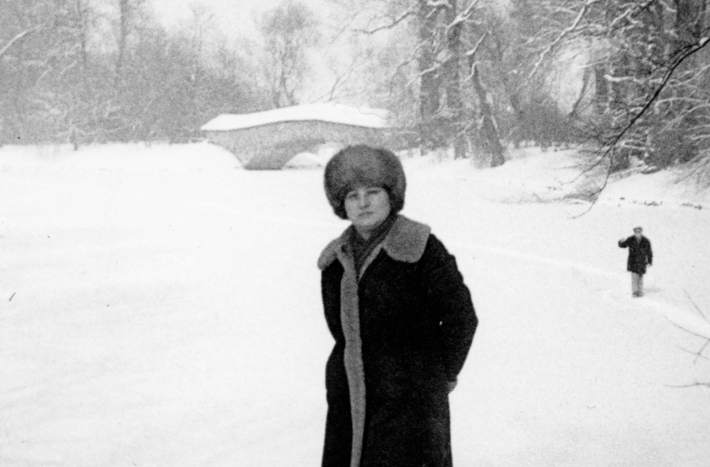 Kobieta w długim kożuchu i puchatej czapce stoi po kolona w śniegu. W tle widać most rzymski i mężczyznę idącego po zamarzniętym jeziorze.