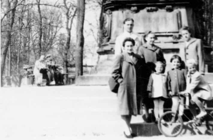 Trzy kobiety, mężczyzna i troje małych dzieci pozują pod pomnikiem. Uśmiechają się. Jedna dziewczynka siedzi na rowerze.