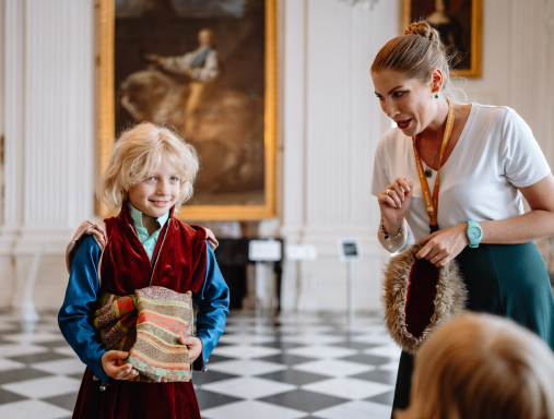 Młoda kobieta stojąca obok chłopca ubranego w strój historyczny. Chłopiec uśmiecha się. Kobieta coś do niego mówi. W głębi ludzie zwiedzający ekspozycję.