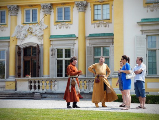 Dwóch mężczyzn ubranych w stroje historyczne. Obok nich ludzie ubrani w stroje współczesne. Grupa stoi przed pałacem w Wilanowie.