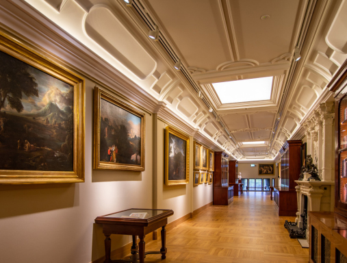 Długi korytarz w pałacu wilanowskim. Na ścianach znajdują się obrazy olejne w złotych ramach. Przy ścianach stoją drewniane gabloty i meble.